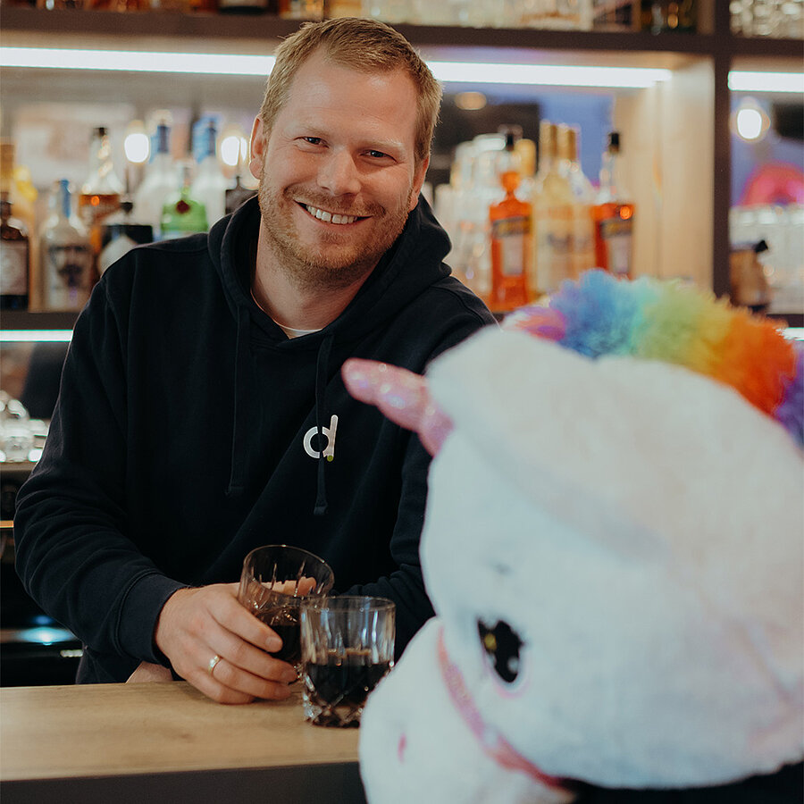 Roland, Projektentwickler bei der B2B Digitalagentur digit.ly in Hannover, trinkt mit einem Plüscheinhorn ein Getränk an der Bar.