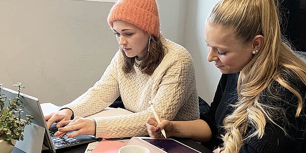 Zwei Mitarbeiterinnen einer Digitalagentur halten eine Besprechung für Social Media Marketing ab. Dabei sitzen sie mit ihrem Laptop an einem Schreibtisch.