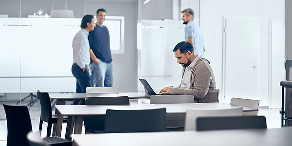 Mitarbeitende der B2B Digitalagentur digit.ly stehen in der Lounge ihres Büros in Hannover, ein Mitarbeiter sitzt am Tisch und arbeitet am Laptop.