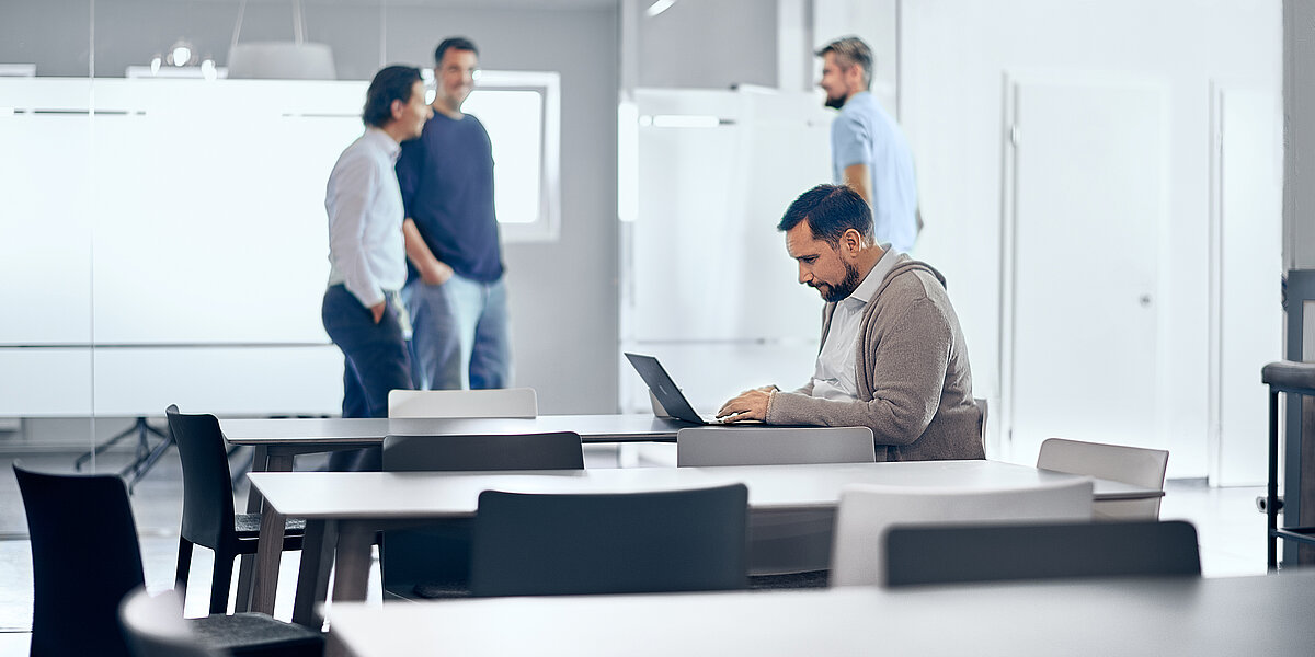 Mitarbeitende der B2B Digitalagentur digit.ly stehen in der Lounge ihres Büros in Hannover, ein Mitarbeiter sitzt am Tisch und arbeitet am Laptop.