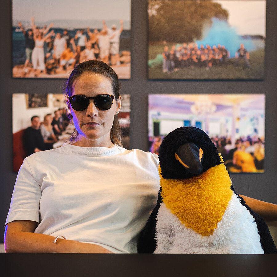 Sarah, HR Managerin bei der digit.ly B2B Digitalagentur aus Hannover, trägt eine Sonnenbrille und sitzt mit ernstem Blick neben einem menschengroßen Pinguin-Kuscheltier auf einem Sofa.